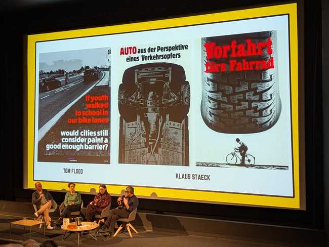 The same two people from the previous pictures are sitting in front of the screen, but now joined by another two
people. The screen is showing black and white and red images of anti-car/pro-cycling advertisements.