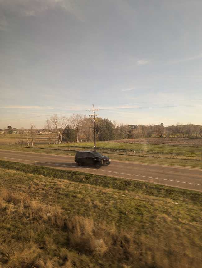 You can see: The verge by the tracks, a road, and then fields with some trees, and distant housing.