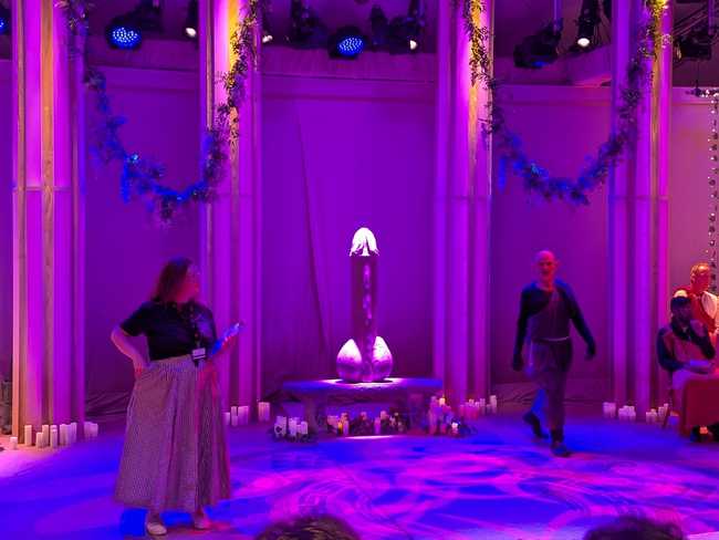 The same stage, with a stage hand on the left, a goblin performer on the right, and a giant erect penis on a bench
under a spotlight.