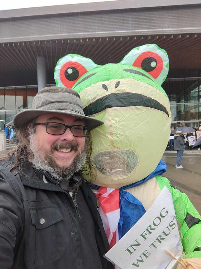A picture of a scruffy soggy man next to someone in an inflated frog costume.
