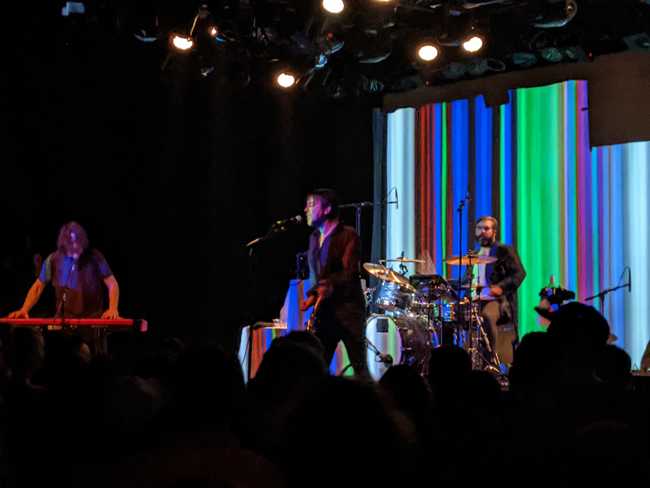 A rock band plays. Crowd silhouette in front, a funky screen behind
them.