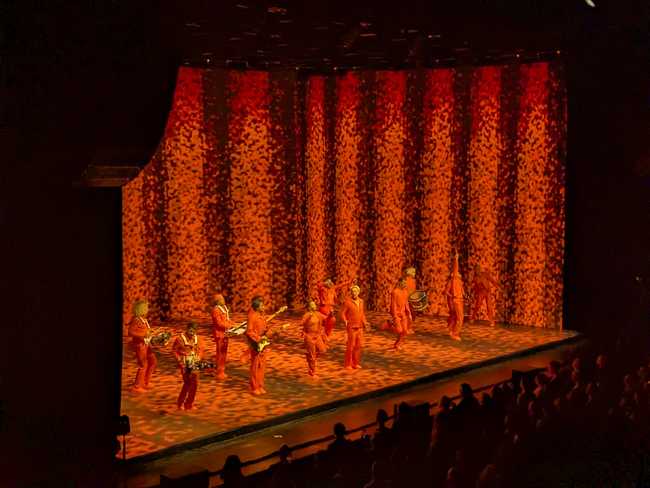 The walls and floor are filled with orange particles against black. The performers not holding instruments are jumping
wildly.