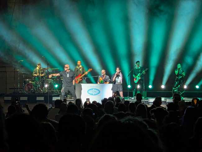 Aqua on stage. Blue and green spotlights fan in the background. The foreground is a sea of silhouetted audience heads.