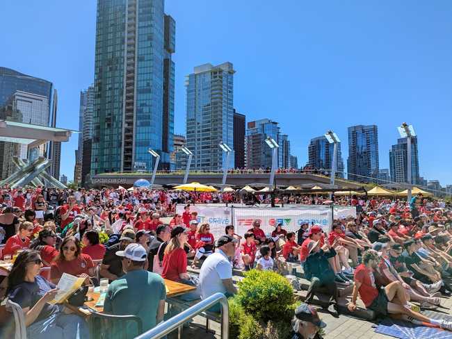 A crowd of people, mostly wearing red, looking at a screen to the right. You can see Vancouver's Olympic Cauldron in
the background, and the skyline of Coal Harbour.