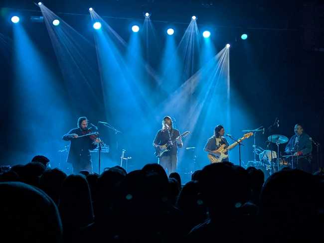 Concert picture. From left to right, there is a man playing the violin, another playing a twelve-string guitar and
singing, a guy on bass, and the drummer.