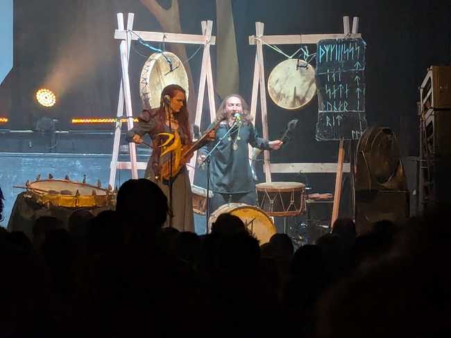 A woman plays some sort of stringed instrument while a man drums and sings.