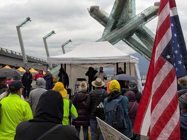 A group of people in the rain listening to a man speaking under a tent. There's an upside down American flag on the
right.