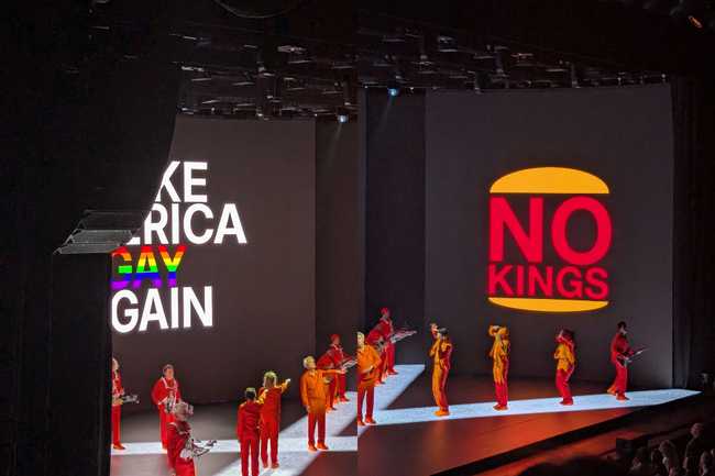 On the left, you can see one screen with the obscured slogan "Make American Gay Again" ("gay" is rainbow-coloured). On
the right, you can see a Burger King logo reading "No Kings". The performers on stage are standing in a cross, with
corresponding cross bars on the floor. The connection between the left and right sides is disjointed.