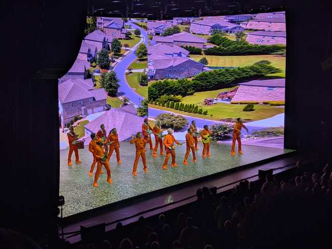 Concert. The walls show an American suburb from above, with generic big houses on a windy street. The floor looks like
astroturf. Everyone is wearing an orange jump suit.