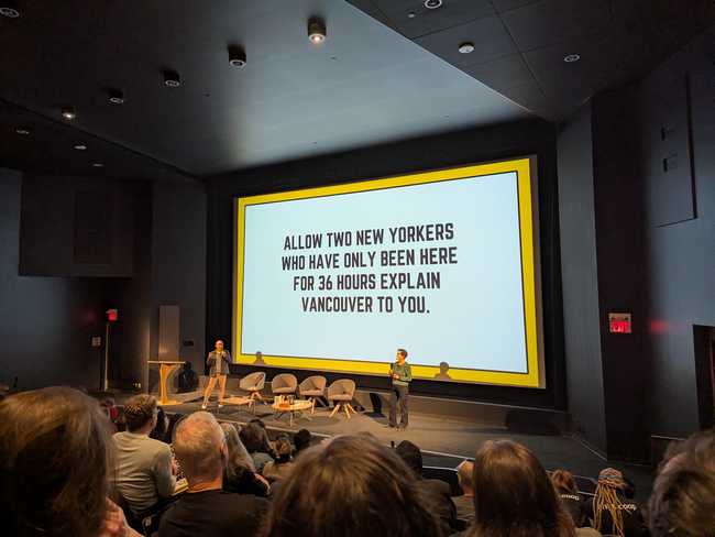 A further out view of the same two people in front of a screen. It displays the text "Allow two New Yorkers who have
only been here for 36 hours explain Vancouver to you." You can see some of the crowd watching.