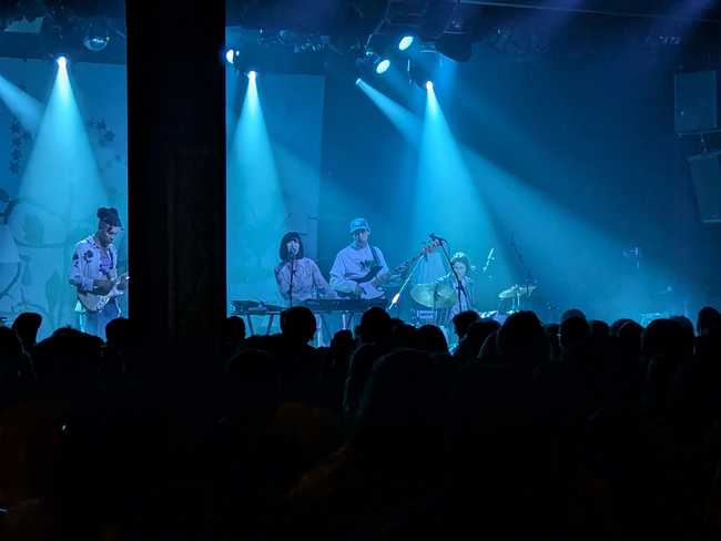 Four people on tag in front of blue flood lights. From left to right, there's a guitarist on the far left, then a
shorter woman on keyboard singing into a microphone, a guy playing bass guitar, and the
drummer.