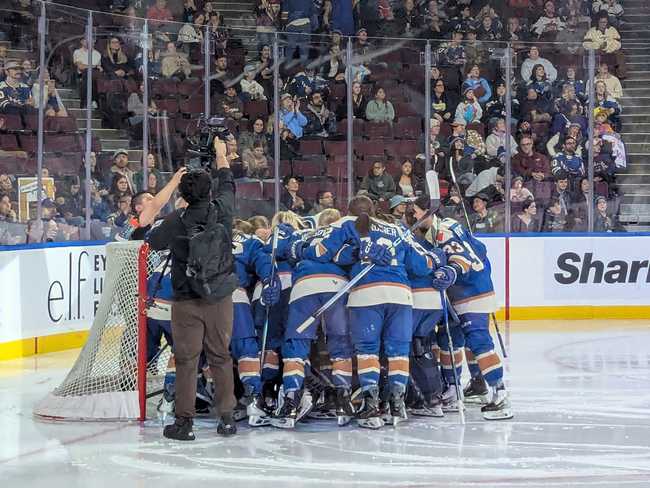 A crowd of hockey players huddle in front of the net.