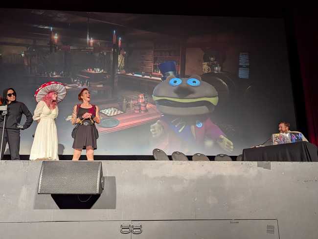 Three performers on the left; the DM at a table on the right. The screen in between shows a fantasy tavern. There's a
guy in fancy dress with a burger for a head looking at the audience.