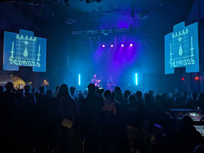 A wide shot showing Devours on stage, the drummer to the left, and the audience. Everything is either blue or black.
