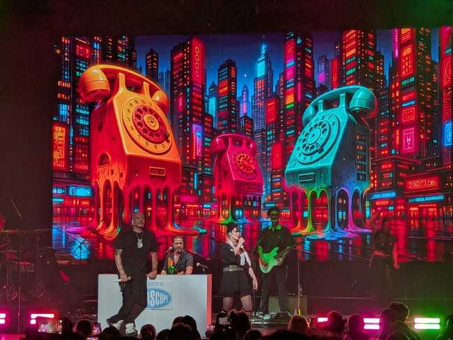Four people on stage. In the background is a neon cityscape with some giant rotary telephones melting into goo. The
numbers on the dials are gibberish.