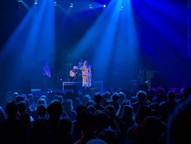 A somewhat distant picture of the show. A woman in a white dress is singing to the crowd. A drummer and bass guitarist
are visible, but obscured by dim lighting.