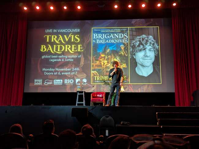 Travis Baldree on stage. The screen behind him has a blurb for the event, the book cover for Brigands & Breadknives,
and his author photo.