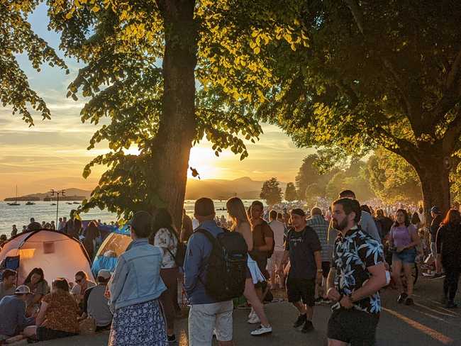 A large group of people are packed along the shore. The sunset gives
it a golden glow.