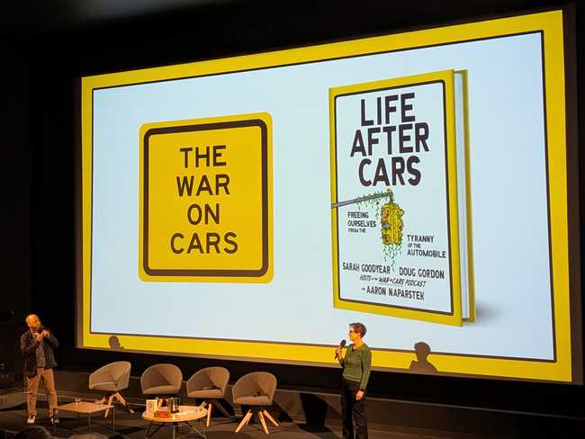 Two people in front of a large screen. On the left is a what looks like a yellow traffic sign with the text "The War
on Cars". On the right is a hardcover book.