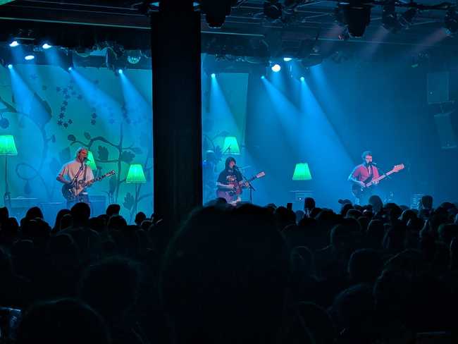 A different band on the same stage. You can see a guy on guitar on the left, a woman on guitar in the middle, and a
guy on bass on the right. They're all singing into microphones. In the back you can faintly see the drummer behind an
inconvenient pillar.
